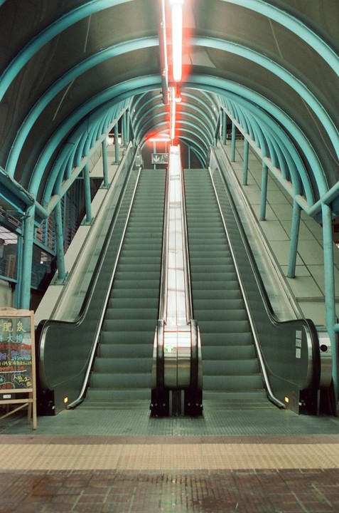 Two escalators ascend under a blue arched tunnel with a red neon light.