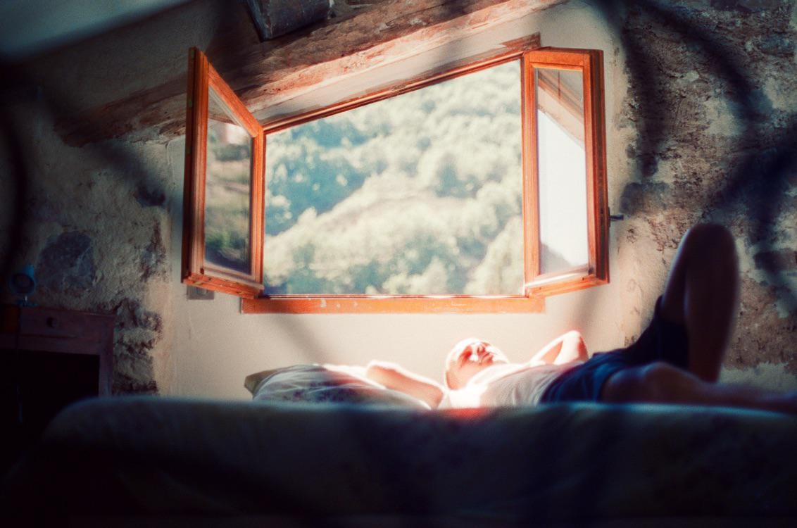 A man lies on a bed in a sunlit room with an open window looking out onto a forest.
