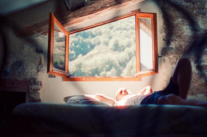 A man lies on a bed in a sunlit room with an open window looking out onto a forest.