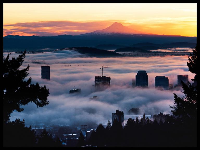 A city skyline is covered in fog with a mountain and colorful sky in the background.