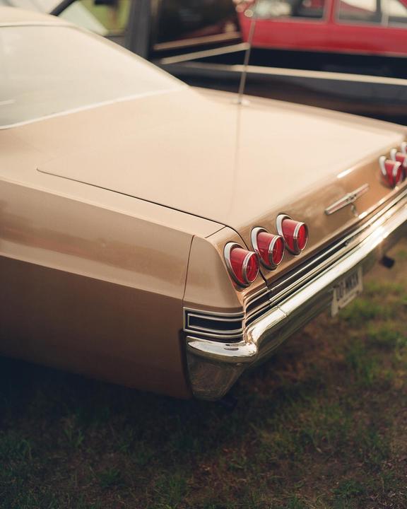 The rear of a vintage tan car with three round red taillights is parked on grass.