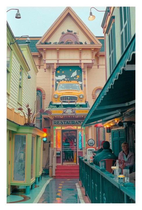 A yellow car is mounted on the facade of a restaurant with neon signs and people sitting at tables outside.