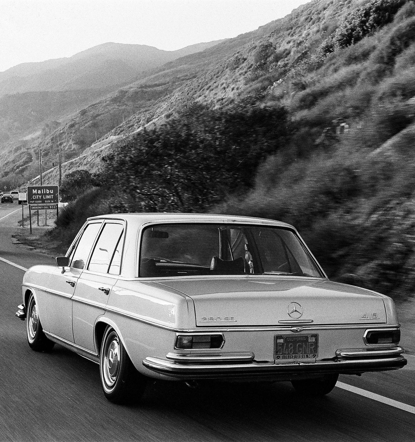 A car drives on a highway next to a Malibu city limit sign with mountains in the background.