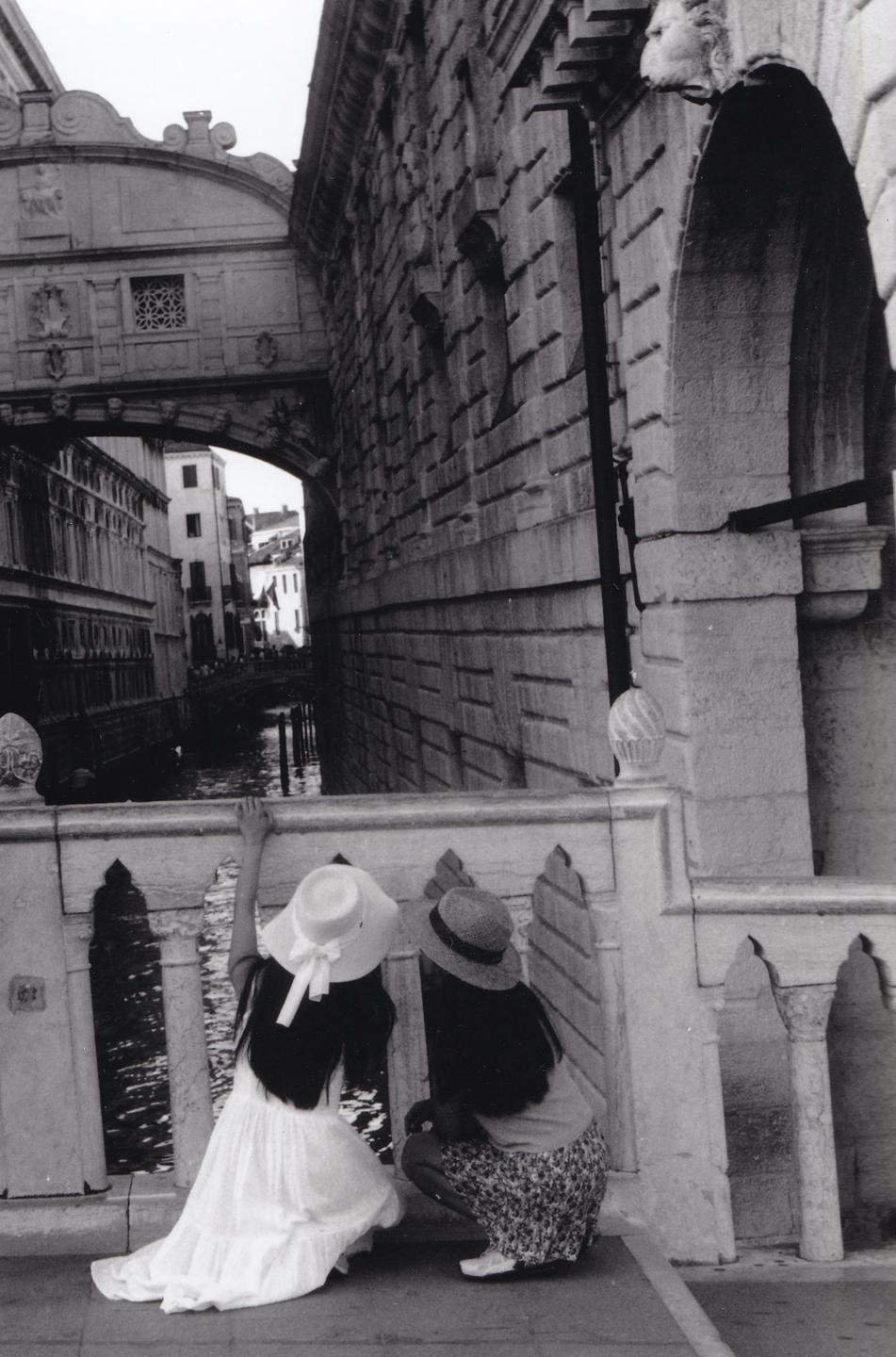Two women wearing hats look over a stone bridge at a canal in Venice.