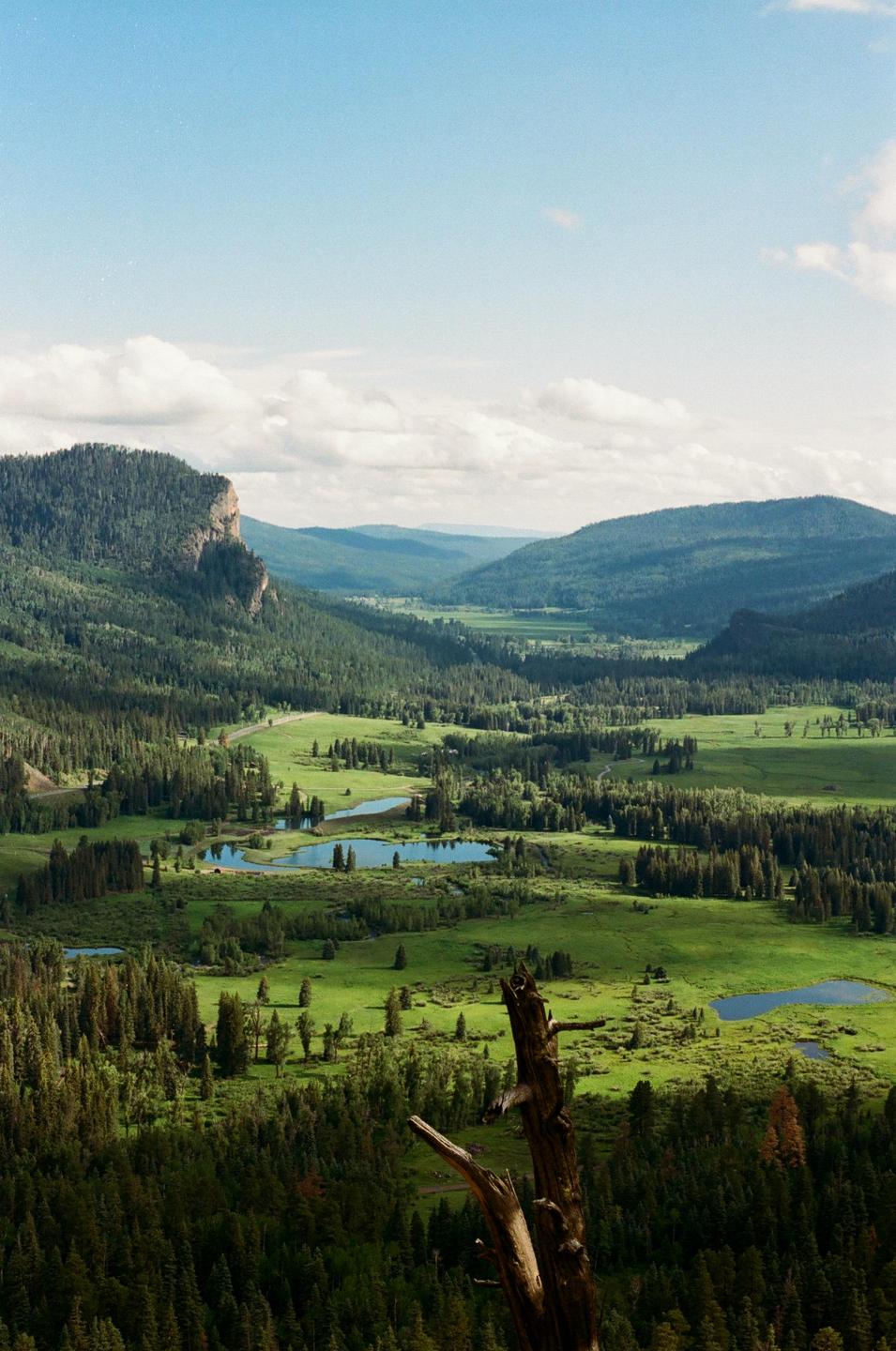 A vast valley with green meadows, forests, and several small lakes is visible under a partly cloudy sky.