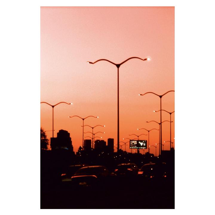 A city street at dusk is filled with cars and streetlights, silhouetted against an orange sky.