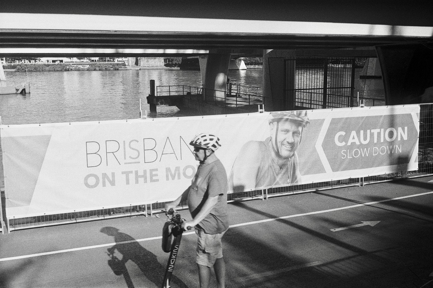 A man in a helmet stands with an electric scooter next to a banner that reads "Brisbane on the Mo" and "Caution Slow Down" under a bridge.