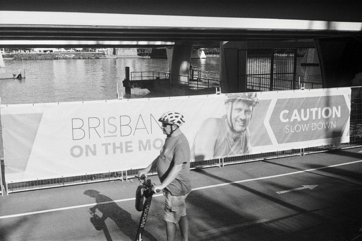 A man in a helmet stands with an electric scooter next to a banner that reads "Brisbane on the Mo" and "Caution Slow Down" under a bridge.