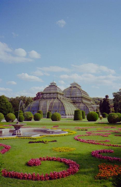 A large glass greenhouse stands behind a fountain and a manicured lawn with flowerbeds.