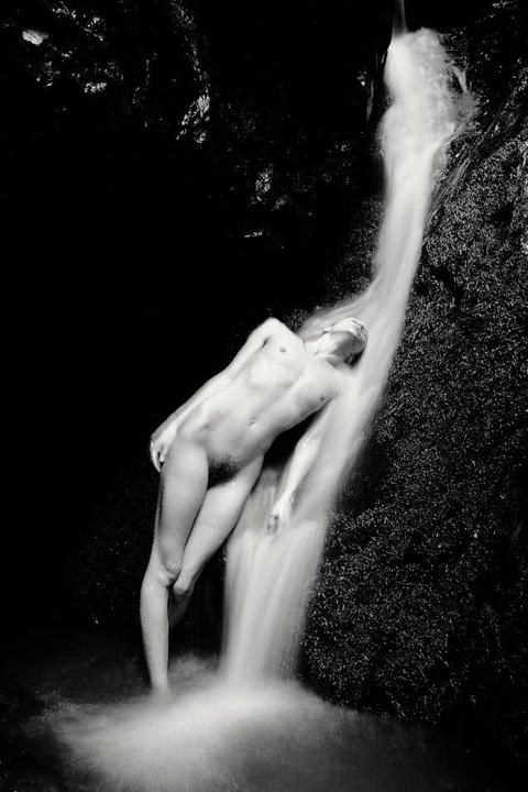 A nude man stands under a waterfall in a dark grotto.