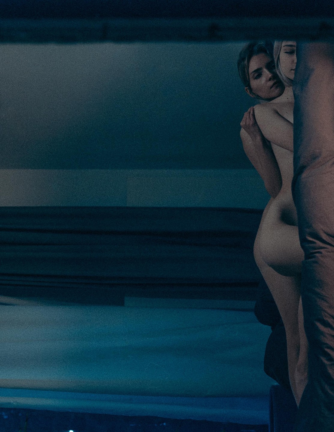 Two women embrace in a dimly lit room, with one woman's back to the viewer and the other looking over her shoulder.