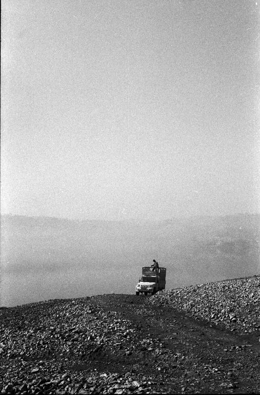 A man sits on the back of a truck driving up a rocky hill.