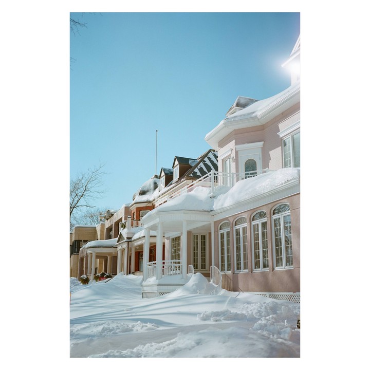 A row of houses with snow-covered roofs and porches line a street under a clear blue sky.