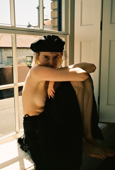 A woman wearing a black hat with a veil sits by a window, looking directly at the viewer.