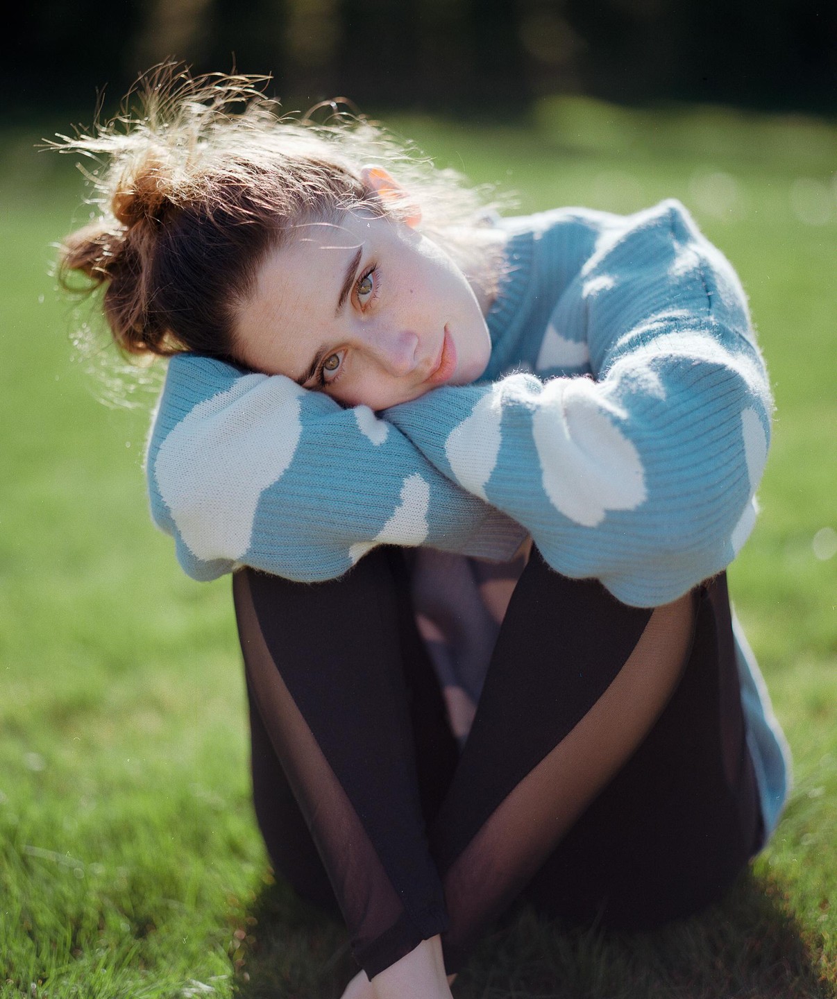 A woman wearing a blue and white sweater sits on green grass with her head resting on her arms.
