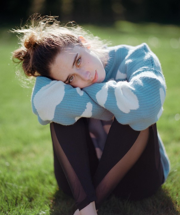 A woman wearing a blue and white sweater sits on green grass with her head resting on her arms.
