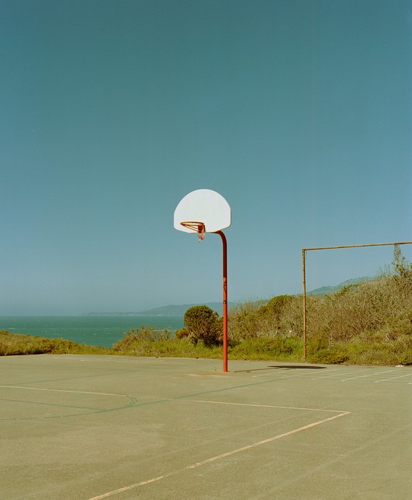 A basketball court with a single hoop stands near the ocean under a clear sky.
