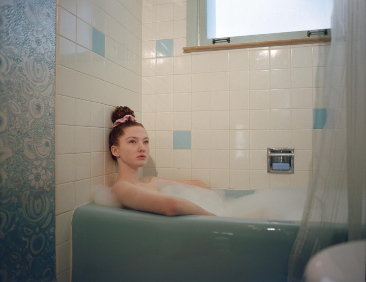 A young woman with red hair in a bun takes a bubble bath in a blue bathtub.