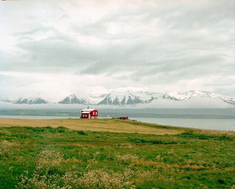 A red house sits on a grassy hill overlooking a body of water with snow-capped mountains in the background under a cloudy sky.
