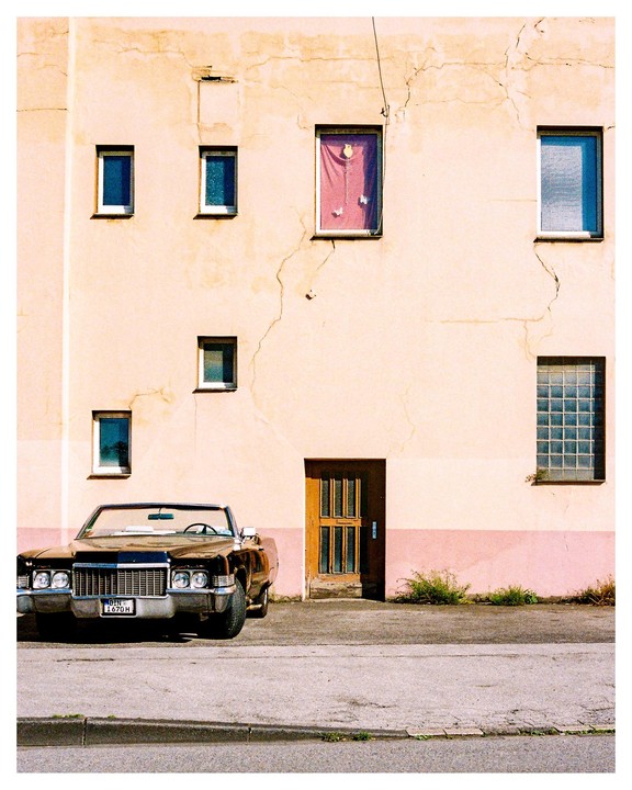 A brown convertible car is parked in front of a building with a pink and beige facade.
