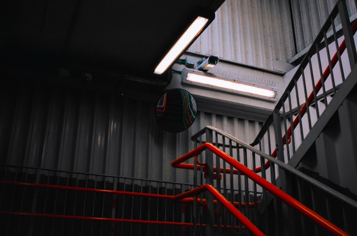 A dark stairwell with red railings and a convex mirror reflecting neon lights.