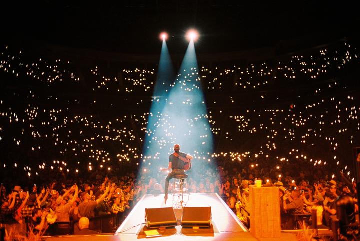 A musician sits on a stool on a stage, playing a guitar under two spotlights, facing a large audience holding up lights.