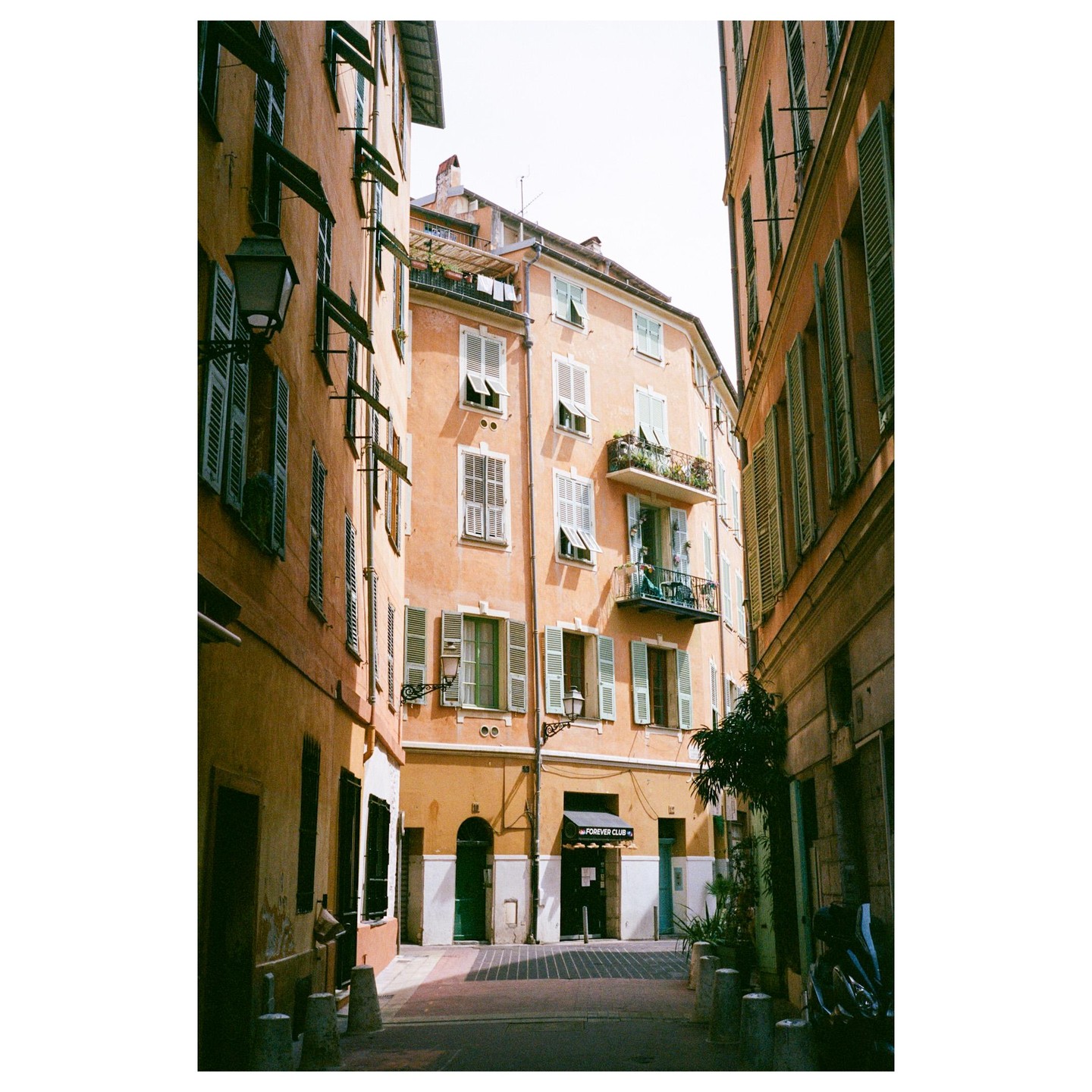 A narrow street is lined with tall buildings featuring many windows with open shutters and a small balcony with plants.