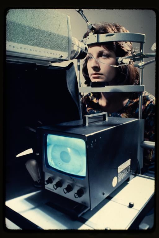 A woman is looking into an eye-tracking device with a television camera and a monitor displaying an eye.