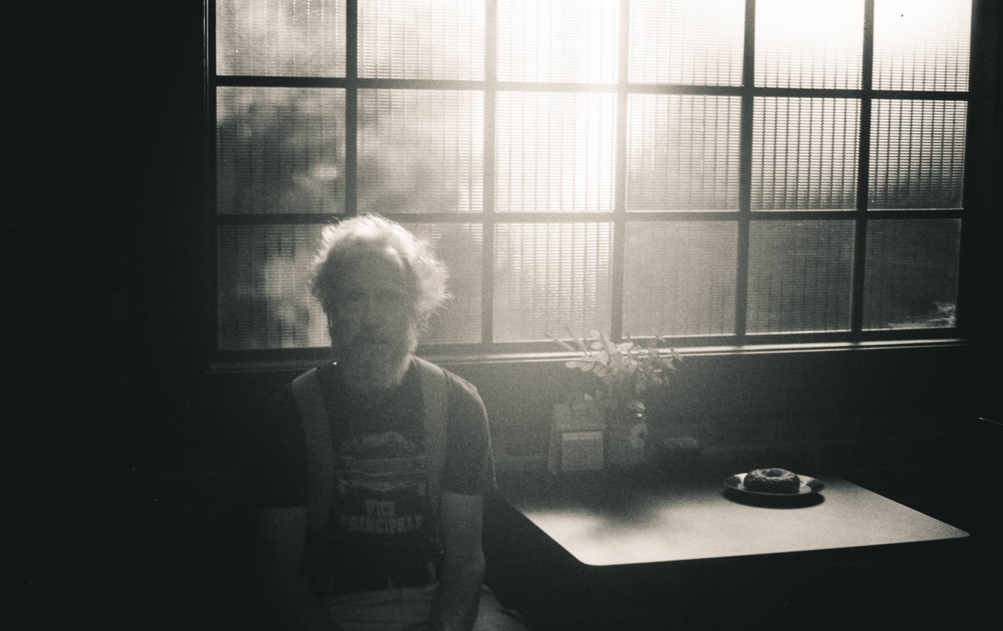 A man with a beard sits at a table in a dimly lit room with a window behind him, next to a plate with a donut.