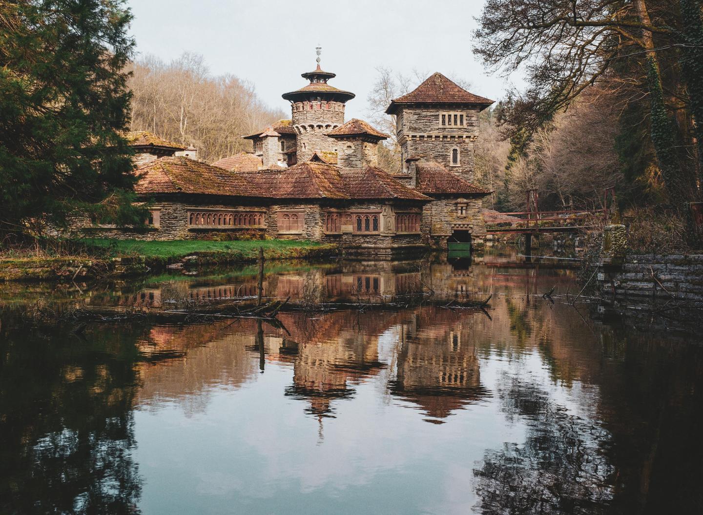 A stone building with a red tile roof is reflected in the still water of a lake, surrounded by trees.