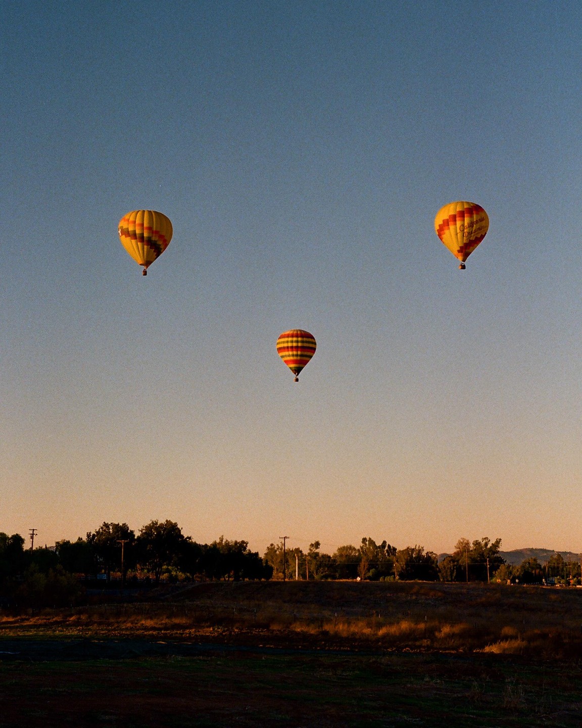 Three hot air balloons float in a clear sky above a rural landscape at sunset.