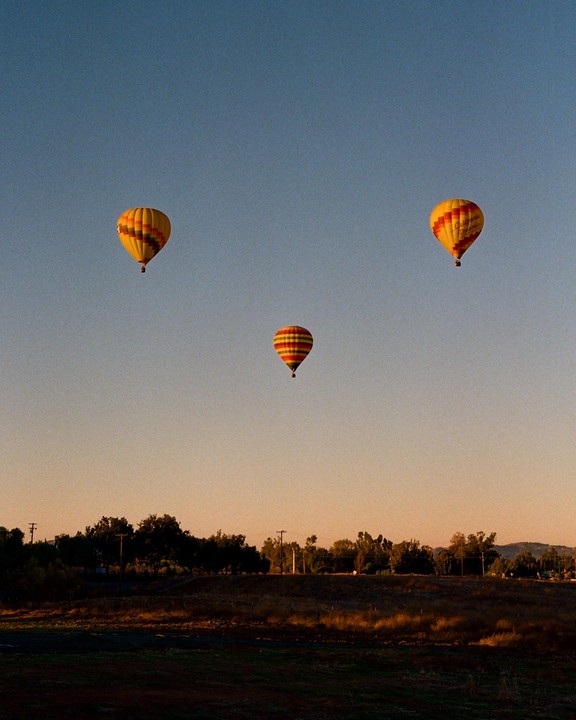 Three hot air balloons float in a clear sky above a rural landscape at sunset.