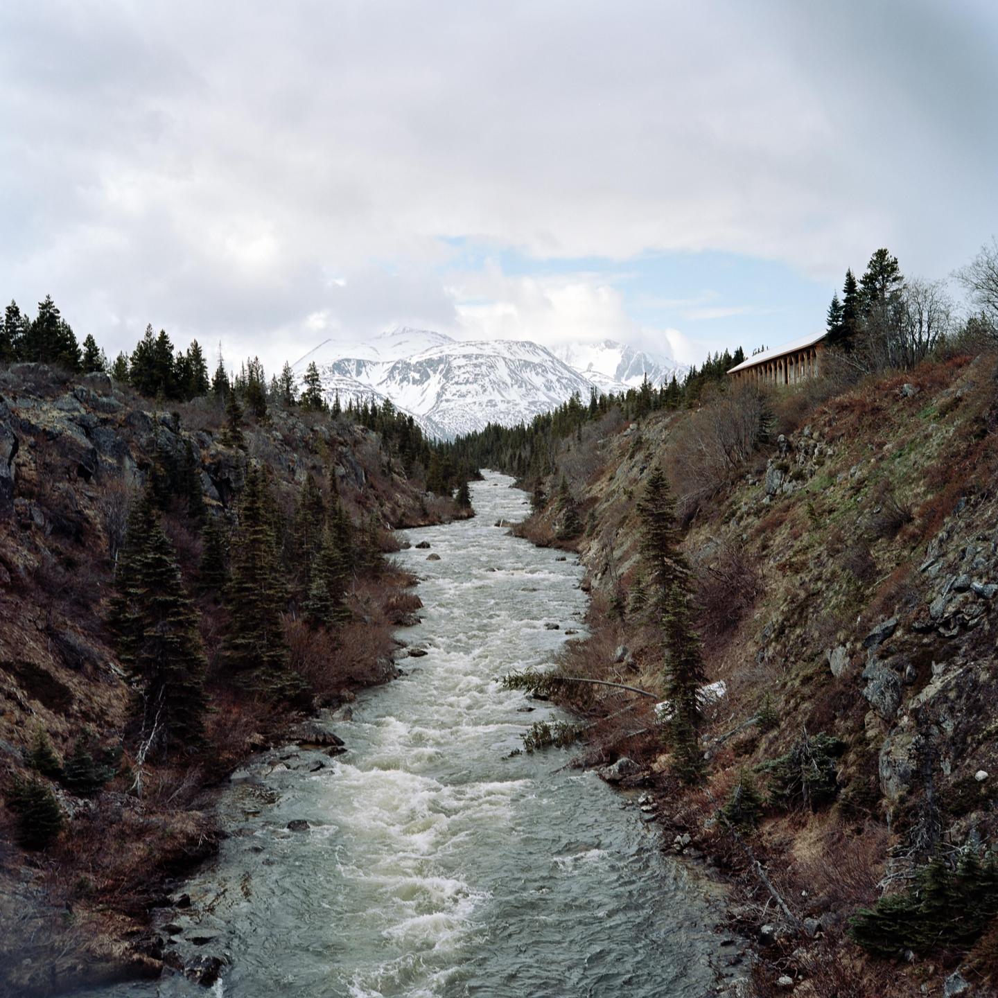 A river flows through a valley with snow-capped mountains in the distance and a building on the right hillside.