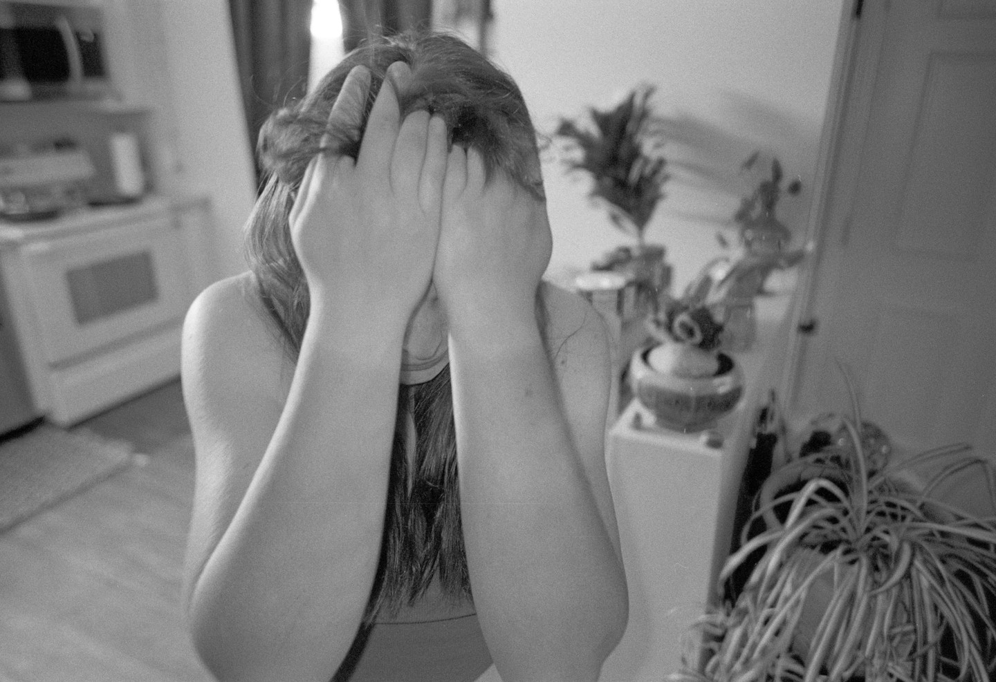 A woman covers her face with her hands in a room with plants and an oven.