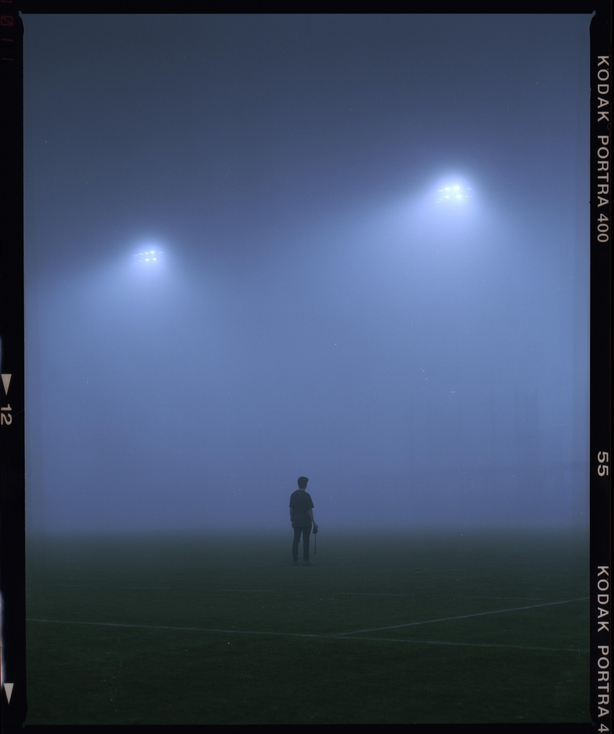A lone man stands on a foggy sports field at night, illuminated by two bright stadium lights.