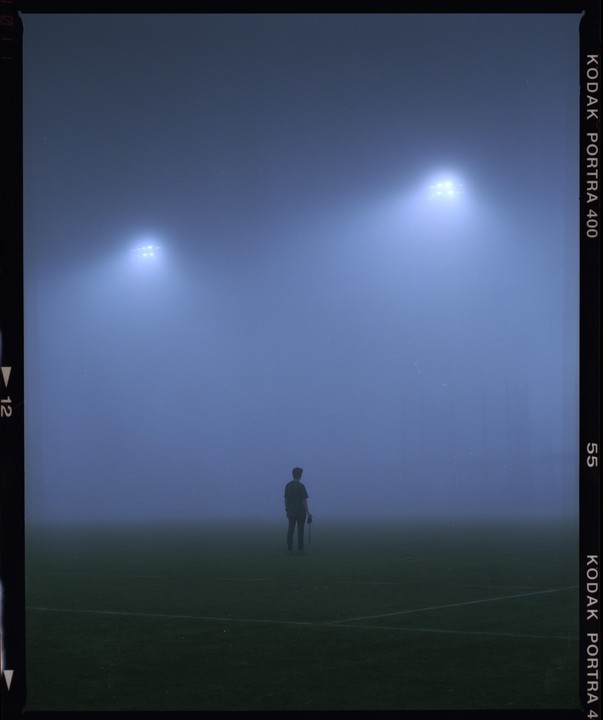 A lone man stands on a foggy sports field at night, illuminated by two bright stadium lights.