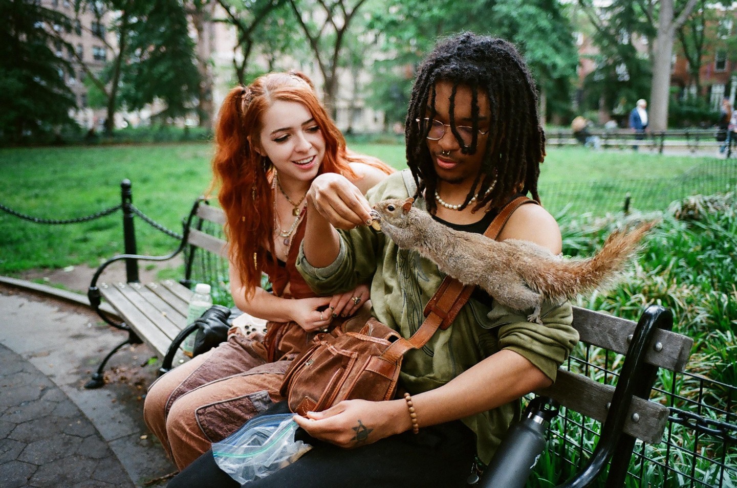 A man feeds a squirrel on his shoulder while a woman watches him on a park bench.