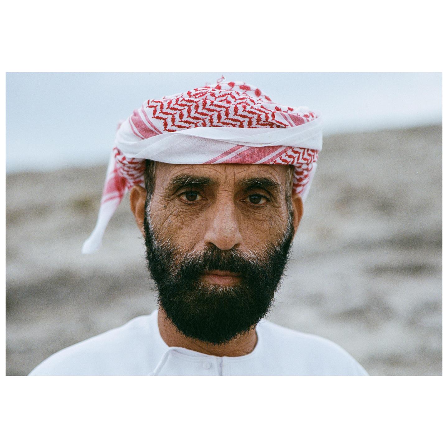 A man with a beard and a red and white head covering looks directly at the viewer.