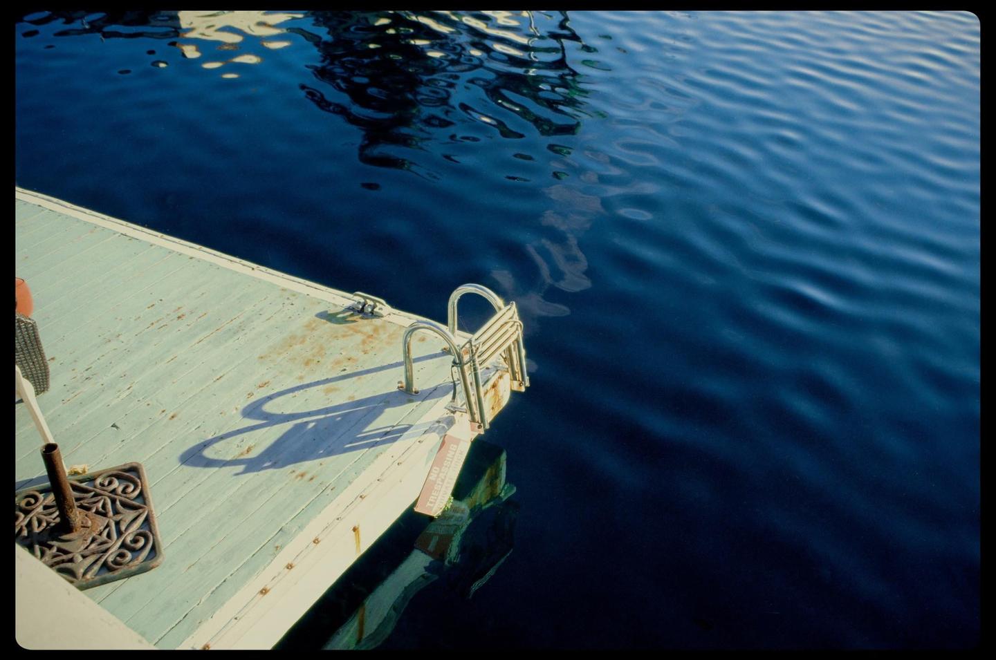 A wooden dock with a metal ladder extends into deep blue water with reflections.