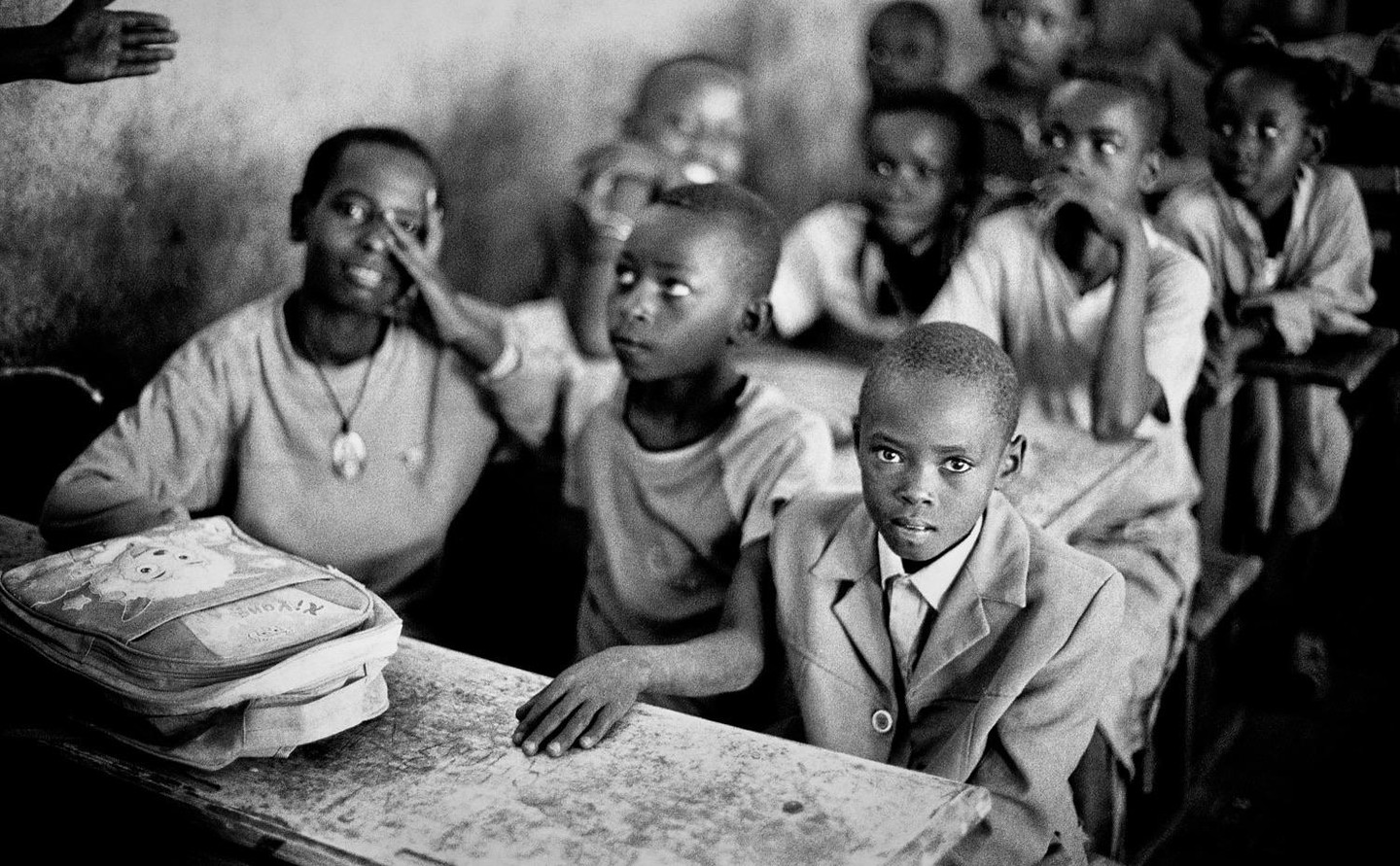 A group of children are sitting at desks in a classroom, with one boy in the foreground looking directly at the viewer.