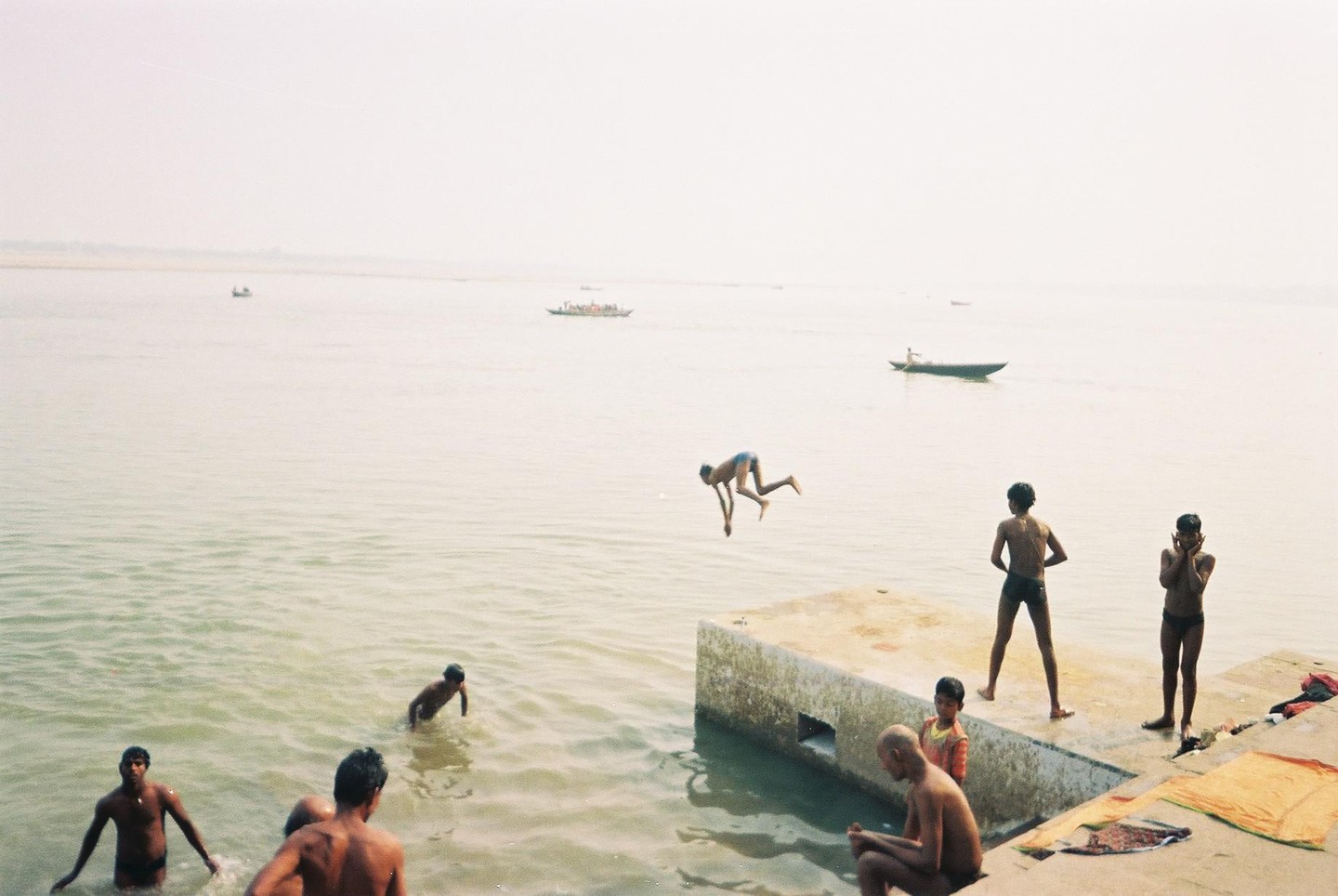 A boy jumps into a river while other boys and men are in the water or on the concrete bank.