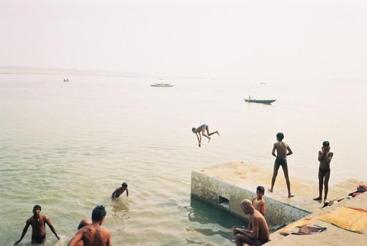 A boy jumps into a river while other boys and men are in the water or on the concrete bank.