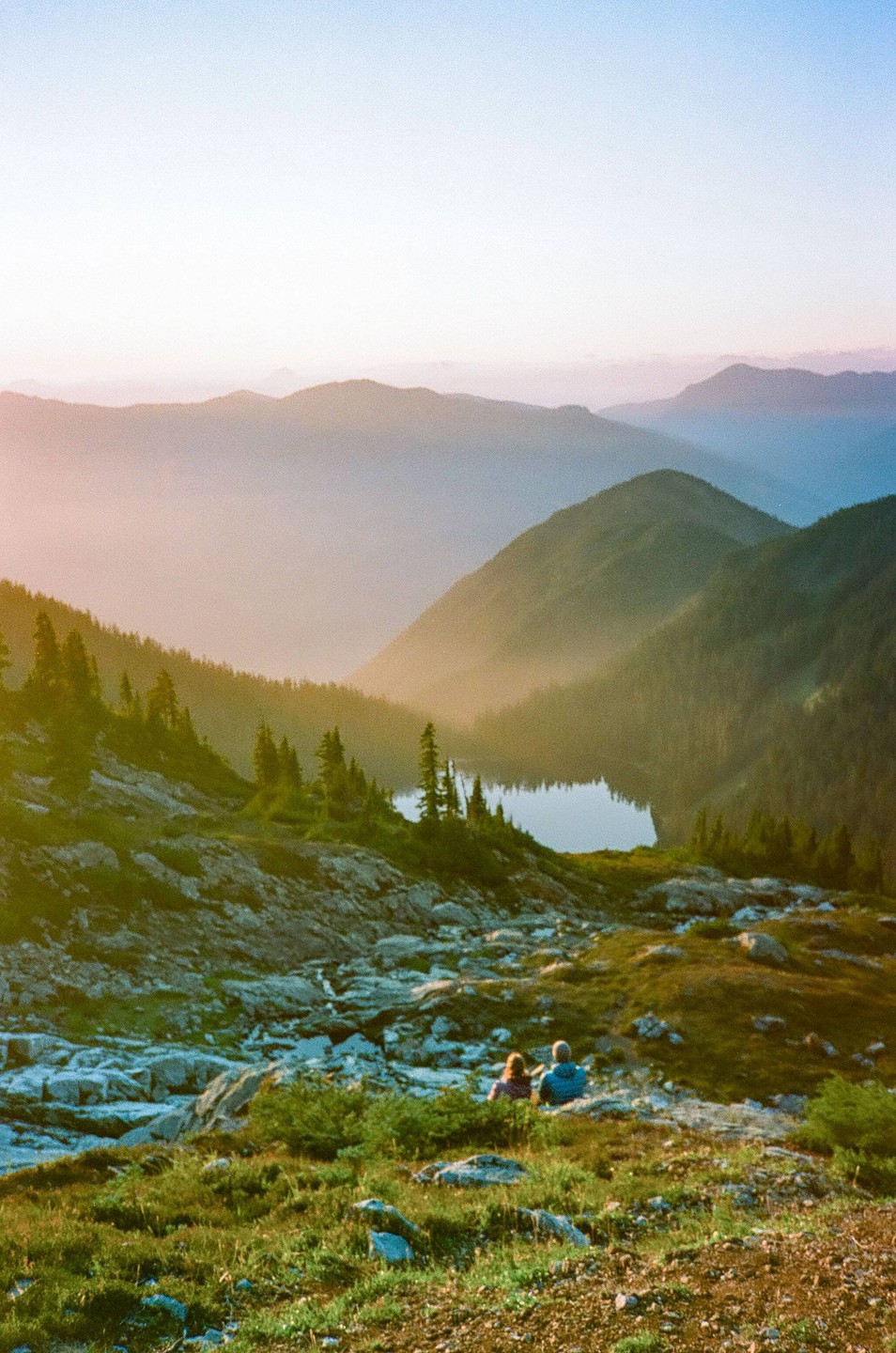 A couple sits on a grassy mountain slope overlooking a lake surrounded by mountains at sunset.