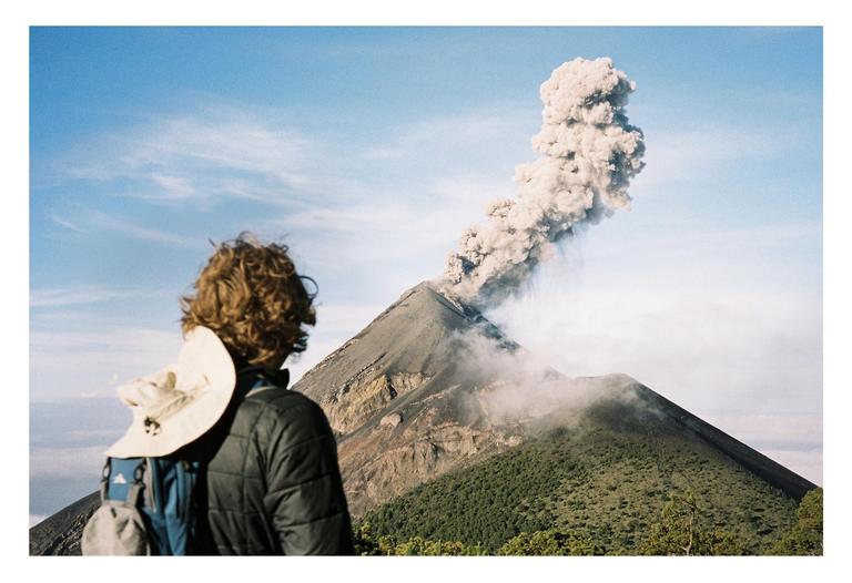 A person with curly hair and a backpack looks at a volcano erupting smoke into a blue sky.