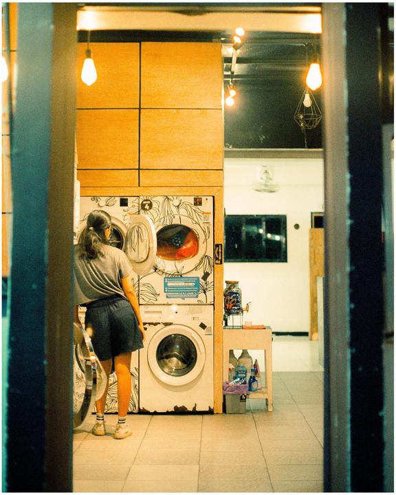A woman stands in front of a washing machine with an open door in a laundromat.