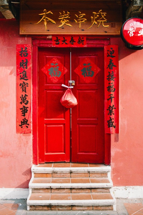 A red door with Chinese characters on banners and a wooden sign above is at the top of a short set of stairs.