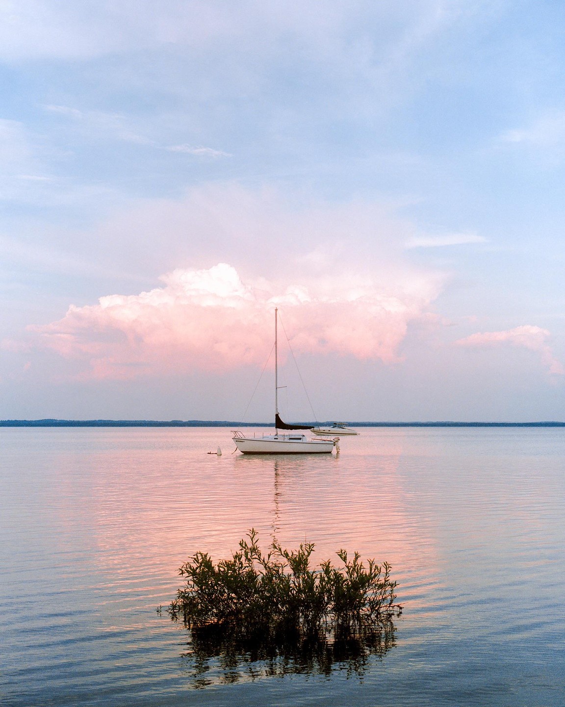 A sailboat floats on a calm lake with pink clouds reflected on the water.