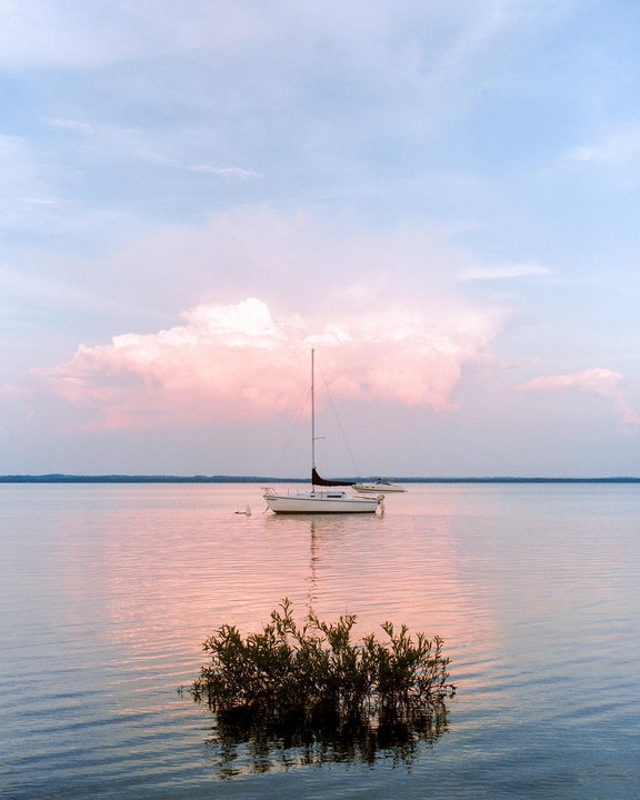 A sailboat floats on a calm lake with pink clouds reflected on the water.