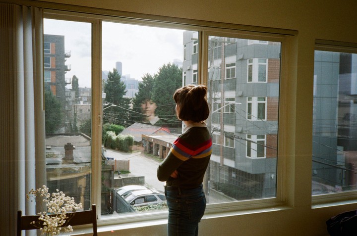 A woman stands with her back to the viewer, looking out a window at a city street and buildings.