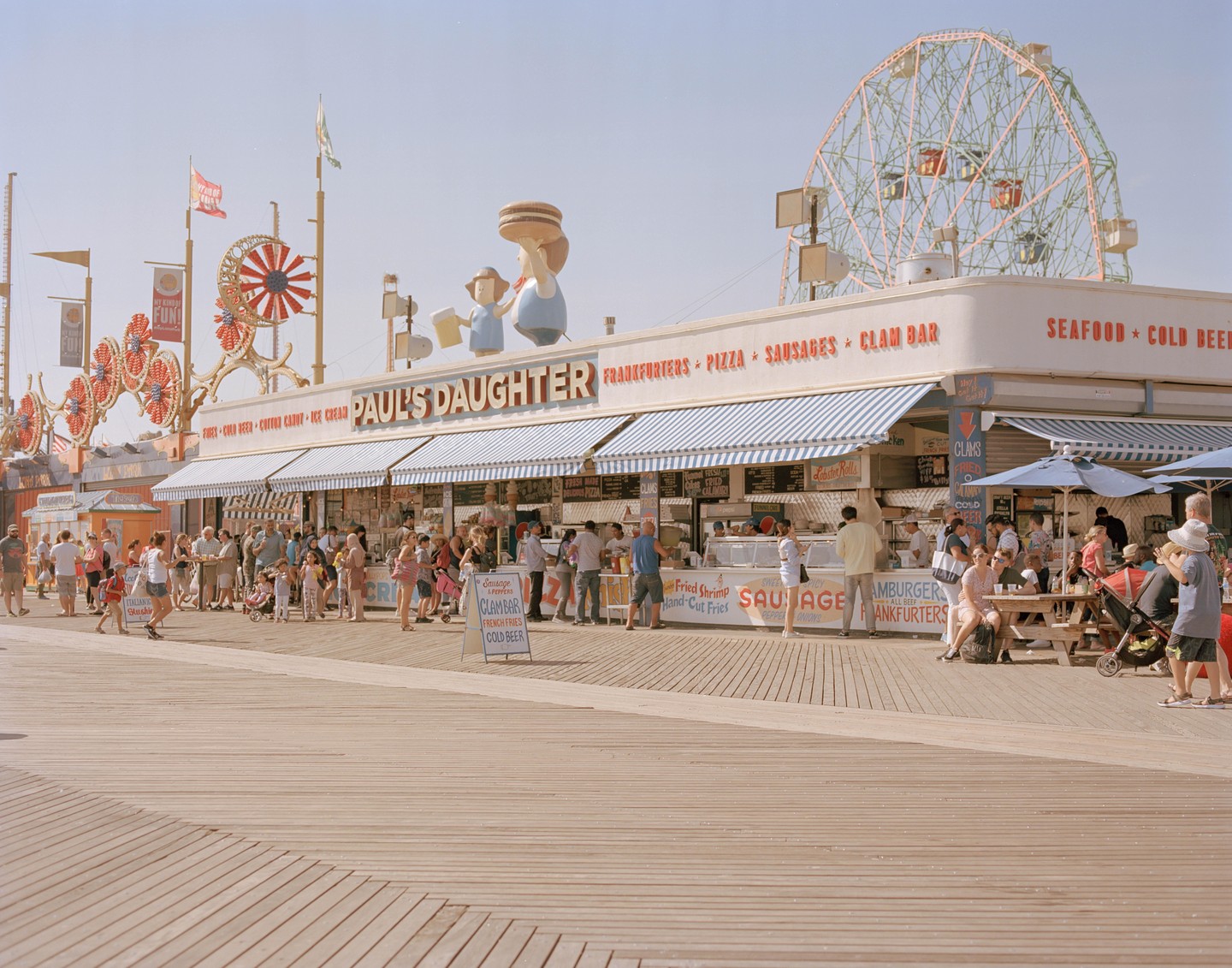 A crowd of people gathers on a boardwalk in front of a food stand with a Ferris wheel in the background.